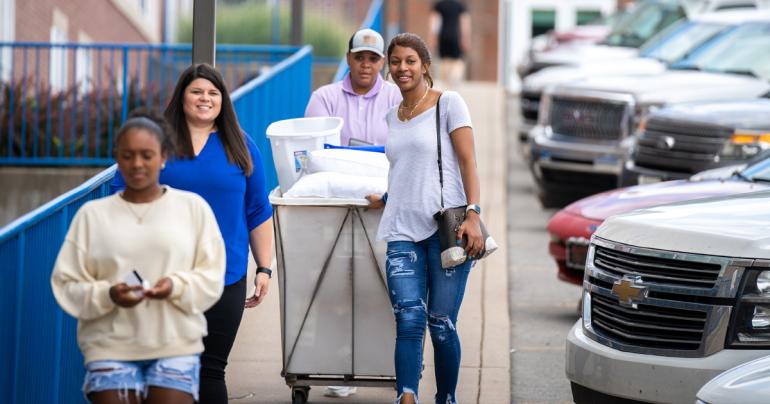 A family wheels a cart of items down the sidewalk in front of Goodwin Hall at Glenville State University as a new student prepares to move in on Monday, August 8. (GSU Photo/Kristen Cosner)